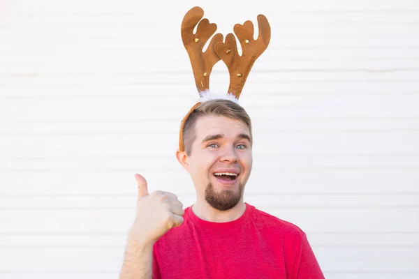 Vacances et Noël concept - Jeune homme aux cornes de cerf avec le pouce vers le haut geste sur fond blanc avec espace de copie — Photo