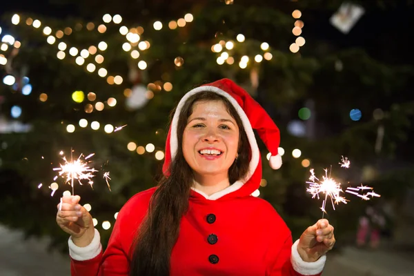 Holiday, Christmas and people concept - Young happy woman wearing Santa suit holding bengal light over Christmas tree background