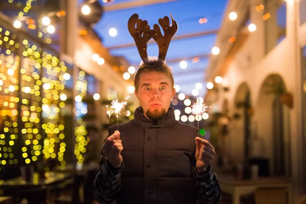 Navidad, gente y el concepto de vacaciones de invierno - hombre sorprendido en cuernos de ciervo de pie en la calle de noche con luces de bengala en sus manos — Foto de Stock