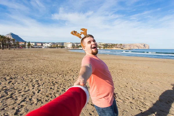 Siga-me, feriados e conceito de Natal - Jovem feliz com chifres de veado segurando a mão do Pai Natal na praia . — Fotografia de Stock