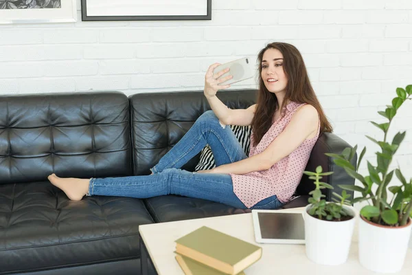 Technology, beauty and people concept - young smiling woman trying to make photo using smartphone and lying on sofa at home touching her hair.