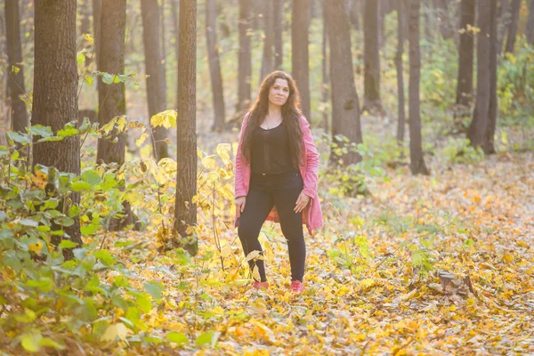 Seizoen, mode en mensen concept- plus grootte vrouw in zwarte broek en roze jas staande in herfst bos — Stockfoto