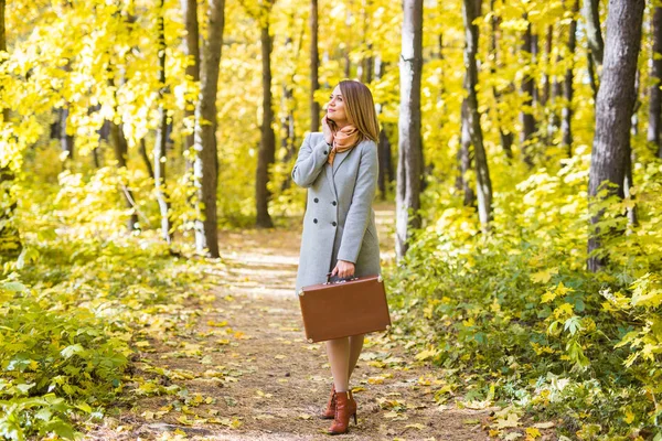 Fashion, season and people concept - happy young woman is going on a trip with retro suitcase on a background of autumn foliage — Stock Photo, Image