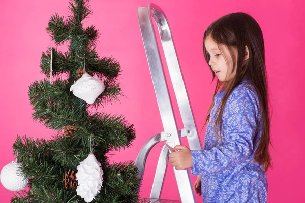 Weihnachts- und Urlaubskonzept - ein kleines Mädchen schmückt Weihnachtsbaum auf rosa Hintergrund — Stockfoto