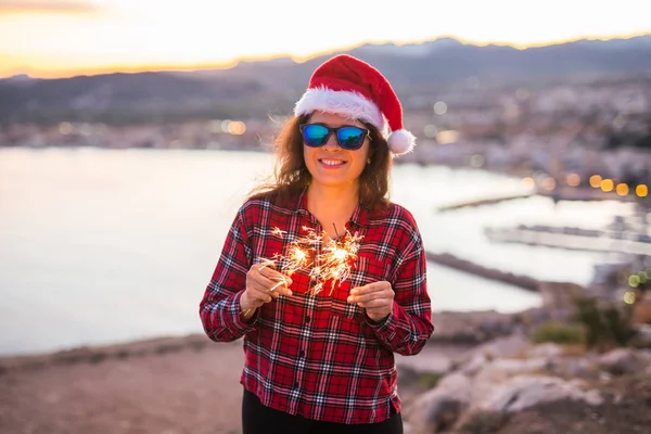 Tatil, Noel ve insanlar kavramı - Noel Baba şapkası havai fişek ve havai fişekleri ile güzel manzara üzerinde genç mutlu kadın