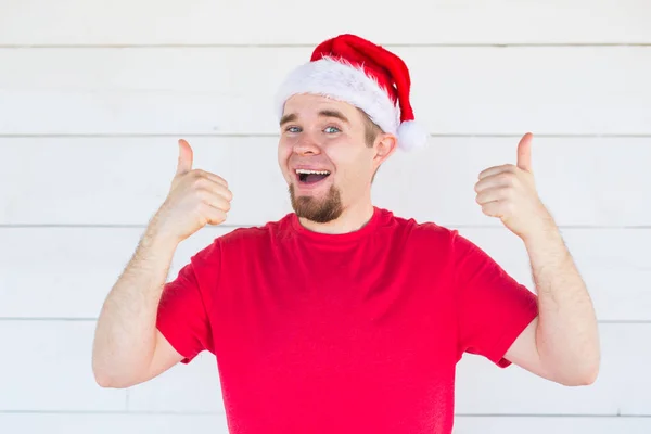 El concepto de la Navidad y la gente - el joven alegre en el sombrero de Navidad que muestra los pulgares hacia arriba sobre el fondo blanco — Foto de Stock
