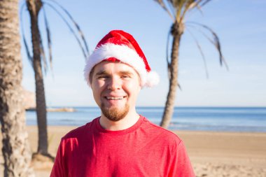 Noel, insanlar, tatil kavramı - plajda gülümseyen Noel Baba şapkası giyen genç adam