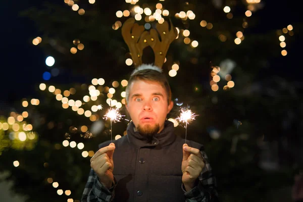 Navidad, gente y el concepto de vacaciones de invierno - hombre sorprendido en cuernos de ciervo de pie en la calle de noche con luces de bengala en sus manos — Foto de Stock