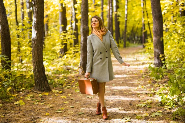 Autumn, fashion, people concept - woman with brown retro suitcase walking through the autumn park and smiling