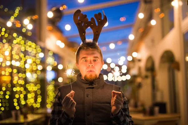 Mensen, vakantie en Kerstmis concept - verrast man in herten hoorns houden twee bengals licht of wonderkaarsen buitenshuis — Stockfoto