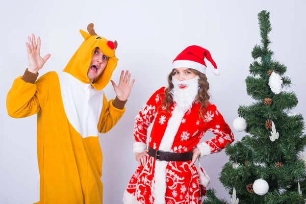 Vacances, Noël et concept de blague - Homme drôle en costume de cerf et femme en costume de Père Noël près de l'arbre de Noël sur fond blanc — Photo