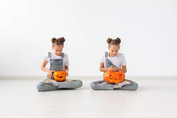 Halloween, bambini e concetto di infanzia - Sorelle gemelle allegre felici sedute con zucche jack-o-lanterna su sfondo bianco — Foto Stock