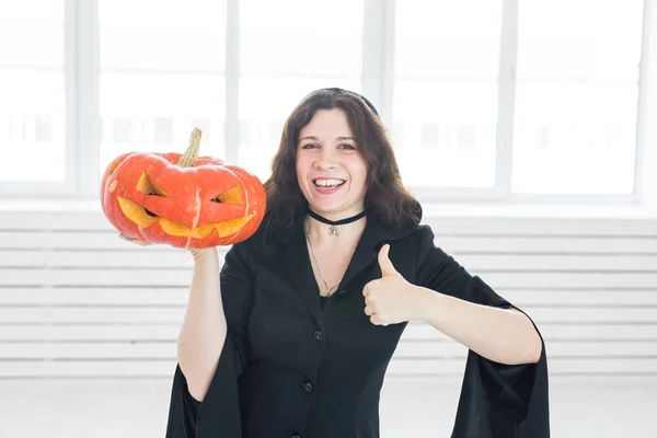 Halloween concept - Happy witch showing thumbs up gesture with pumpkin Jack-o-lantern on light background — Stock Photo, Image