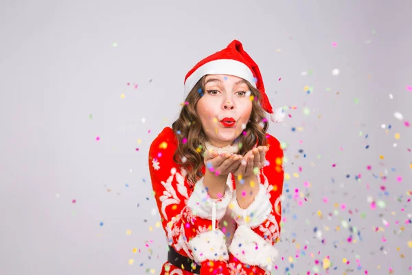 Concepto de vacaciones, Navidad y personas - Mujer hermosa y joven en traje de Santa Claus sobre fondo blanco soplando en confeti — Foto de Stock