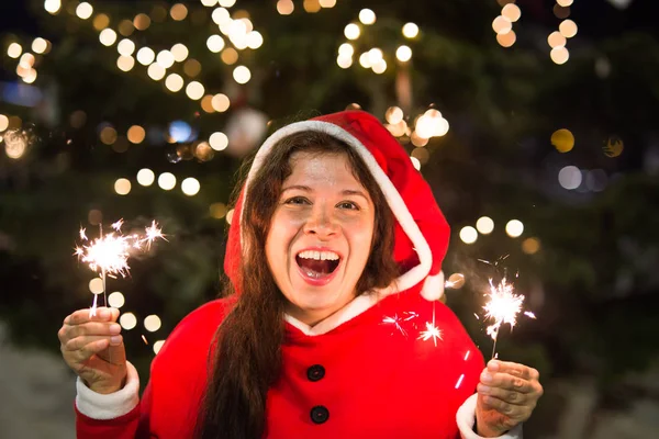 Vacances, Noël et concept de personnes - Jeune femme heureuse portant un costume de Père Noël tenant la lumière du bengale sur le fond de l'arbre de Noël — Photo