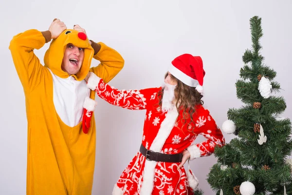 Férias, Conceito de Natal e piada - Homem engraçado em traje de veado e mulher em traje de Papai Noel perto de Árvore de Natal em fundo branco — Fotografia de Stock