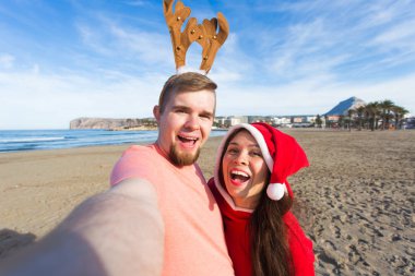 Eğlenceli ve kış tatil kavramı - mutlu çift selfie kumlu plaj arka plan üzerinde alarak Noel kostümleri