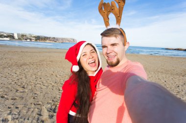 Eğlenceli ve kış tatil kavramı - mutlu çift selfie kumlu plaj arka plan üzerinde alarak Noel kostümleri
