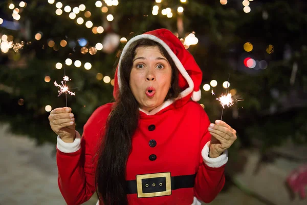 Holiday, Christmas and people concept - Young happy woman wearing Santa suit holding bengal light over Christmas tree background