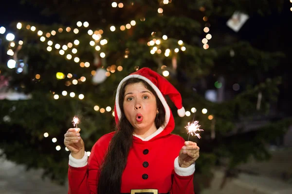 Holiday, Christmas and people concept - Young happy woman wearing Santa suit holding bengal light over Christmas tree background.