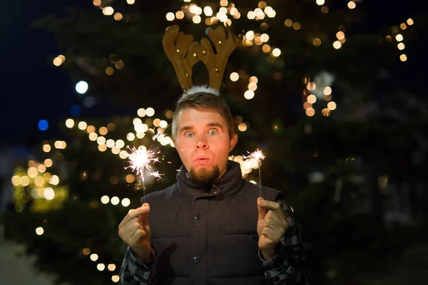 Έννοια διακοπές Χριστουγέννων, άνθρωποι και χειμώνα - έκπληκτος ο άνθρωπος στα κέρατα του ελαφιού στέκεται στο δρόμο το βράδυ με Βεγγάλη φώτα στα χέρια του — Φωτογραφία Αρχείου