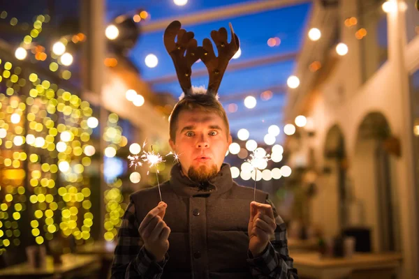 Navidad, gente y el concepto de vacaciones de invierno - hombre sorprendido en cuernos de ciervo de pie en la calle de noche con luces de bengala en sus manos — Foto de Stock