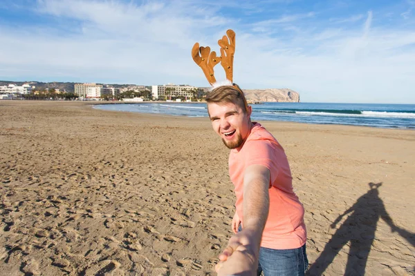 Follow me, holidays and Christmas concept - Happy young man with deer horns holding Santas hand on the beach.