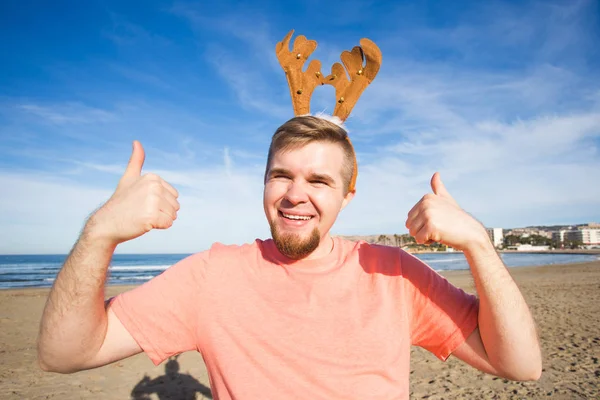 Vacaciones y concepto de Navidad - Hombre con cuernos de rudolph con pulgares en la playa —  Fotos de Stock