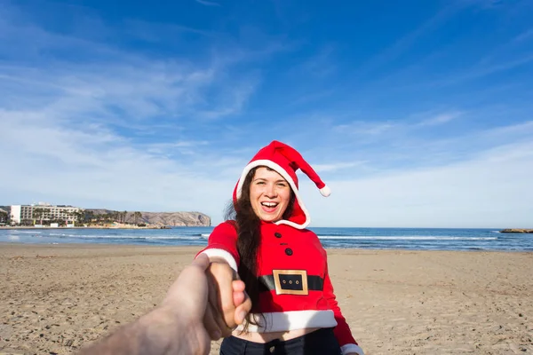 Follow me, holidays and Christmas concept - Happy young woman wearing Santa costume holding hand her friend on the beach.