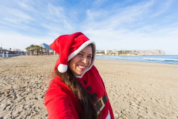 人, 假期和圣诞节的概念-年轻的妇女在圣诞老人服装拍摄自拍在海滩上 — 图库照片