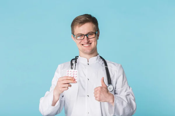 Médico masculino Hansome a mostrar o polegar para cima. Jovem médico segurando pílulas e gesto polegar sobre fundo azul . — Fotografia de Stock