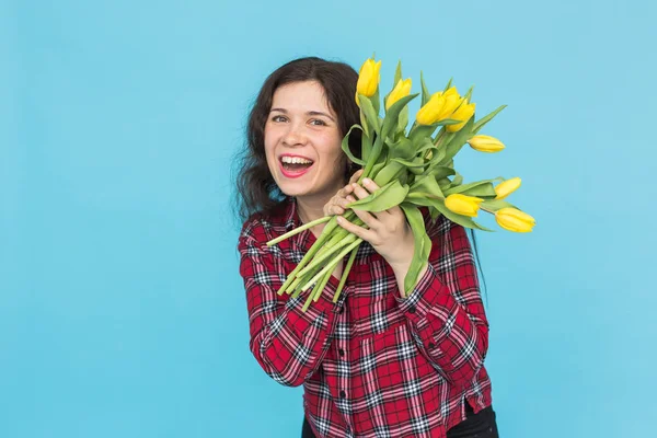 Holidays, flowers and people concept - Funny girl in plaid shirt with bouquet tulips in hands posing on a blue background