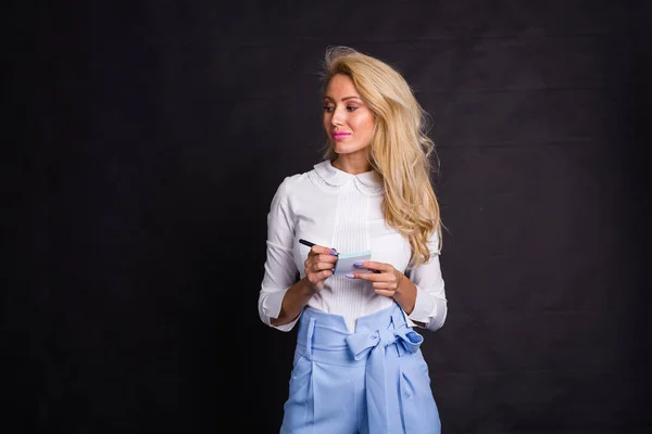 Mode, business en mensen concept - leuke blonde vrouw in spijkerbroek schrijft notities op stickers op zwarte achtergrond met kopie ruimte — Stockfoto