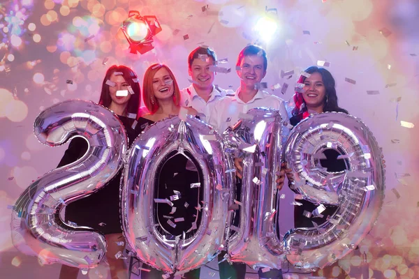 O ano de 2019 está chegando. Grupo de jovens alegres segurando números coloridos de prata e jogando confete na festa . — Fotografia de Stock