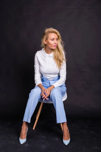 Móda a krása koncept - krásné blond model v bílé košili a džínách, sedí na židli tmavém pozadí — Stock fotografie