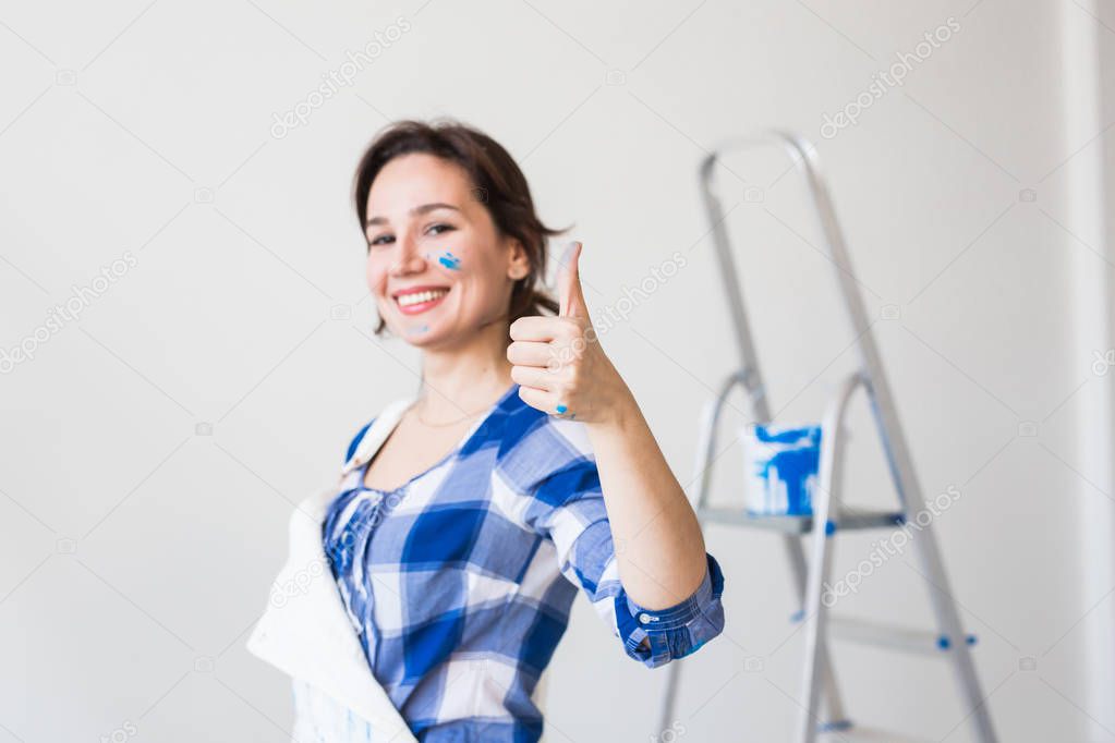 Repair, renovation and people concept - young woman painting the wall and looks like very happy, she showing thumb up