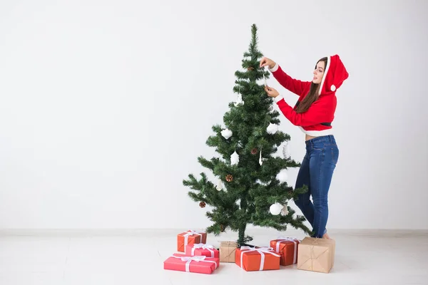 Zimní prázdniny, Vánoce a lidé koncept - šťastná mladá žena oblečená v santa kostým zdobení vánočního stromu v bílém pokoj s kopií prostoru — Stock fotografie