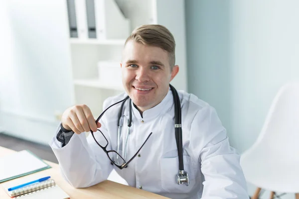 Υγειονομική περίθαλψη, ιατρική και άνθρωποι έννοια - νέος Καυκάσιος γιατρός κάθεται στο γραφείο του και να χαμογελά — Φωτογραφία Αρχείου