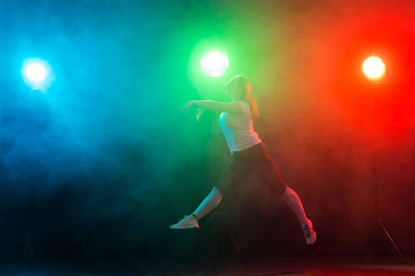 Dancing, sport, jazz funk and people concept - young woman jump in the darkness under colourful light