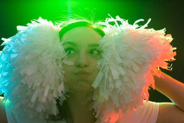 Concepto de baile, animadora y gente: una jovencita divertida en la oscuridad bajo una luz colorida con pompones en la cabeza — Foto de Stock