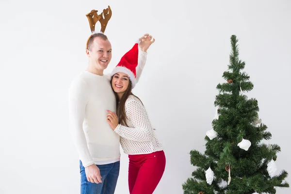 Забавная любящая пара возле рождественской елки. Мужчина носит оленьи рога, а женщина носит шляпу Санты — стоковое фото