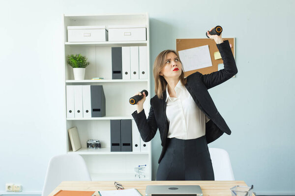 Business, emotions and people concept - Woman in office holding dumbbells in hands