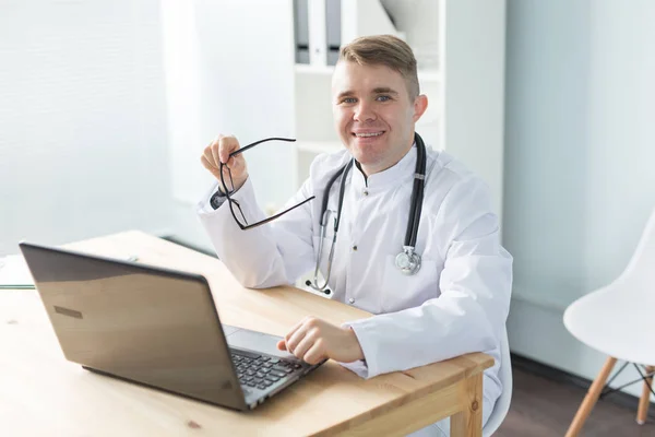 Υγειονομική περίθαλψη, ιατρική και άνθρωποι έννοια - χαμογελώντας ο γιατρός κάθεται στο τραπέζι με το laptop και εξετάζοντας τη φωτογραφική μηχανή — Φωτογραφία Αρχείου