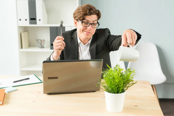 Office, humor, joke and business people concept - handsome man working in office, watering potted plant, sitting with spoon in his hands and smiling.