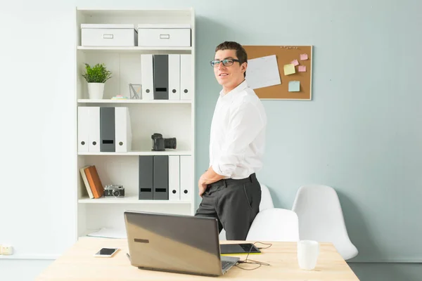 Affärer, kontor, människor koncept - stilig man i glasögon står på kontoret nära en dator och ler — Stockfoto