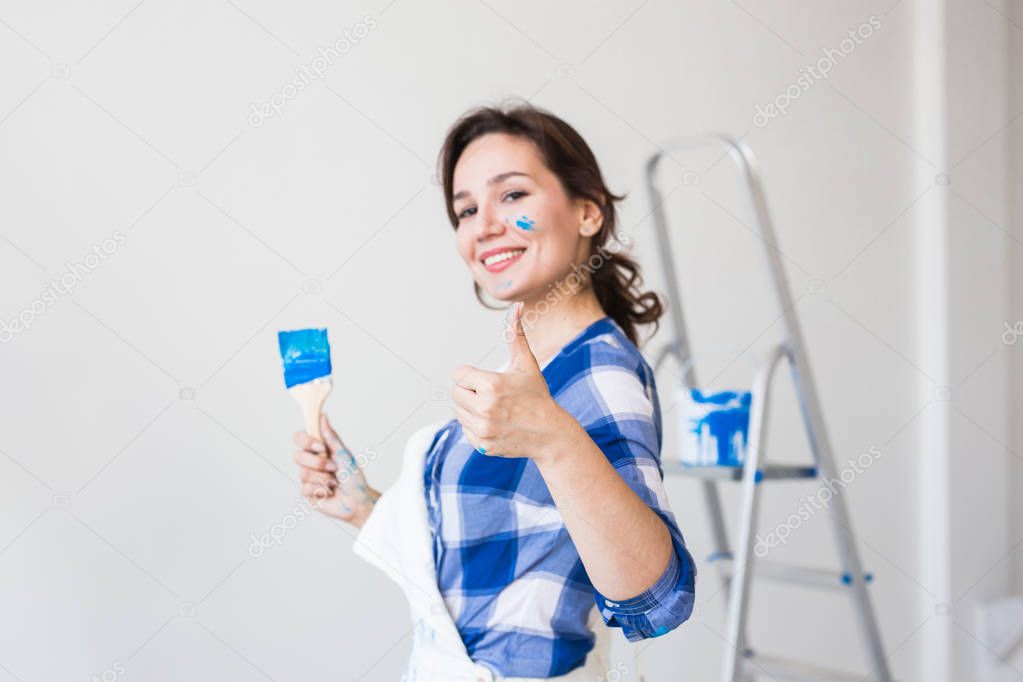 Repair, renovation and people concept - young woman painting the wall and looks like very happy, she showing thumb up