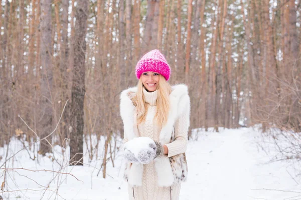 Nature and people concept - Attractive blond woman dressed in coat and pink hat in winter park holding snow in her hands. — Stock Photo, Image