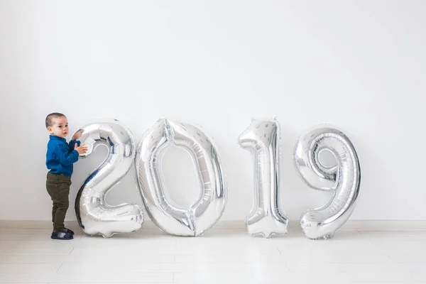 Año nuevo, celebración y concepto de vacaciones - niño pequeño de pie cerca de signo 2019 de globos de plata para el año nuevo en el fondo de la habitación blanca — Foto de Stock