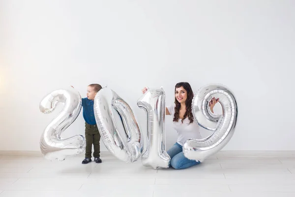 Año nuevo, celebración y concepto de vacaciones - madre e hijo sentados cerca de signo 2019 de globos de plata para el año nuevo en el fondo de la habitación blanca — Foto de Stock