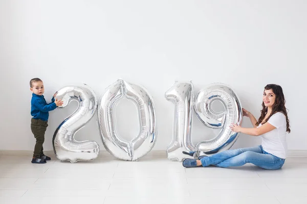 New year, celebration and holidays concept - mother and son sitting near sign 2019 made of silver balloons for new year in white room background